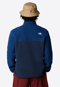 Giacca in pile blu navy con un pannello superiore di un blu più chiaro. Presenta un colletto alto e un dettaglio del logo bianco sul retro. Indossata con un cappello a secchiello color sabbia.