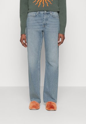 Personne portant un jean droit bleu clair, un pull vert avec un motif de soleil orange, et des chaussons pelucheux orange, debout devant un fond uni.