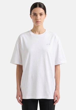 Donna con capelli scuri, indossa una t-shirt bianca oversize a maniche corte e pantaloni neri, in piedi davanti a uno sfondo chiaro e uniforme.