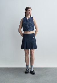 Mujer de pie contra una pared lisa, vistiendo un top corto azul sin mangas con botones, falda plisada azul marino, calcetines grises y mocasines negros.