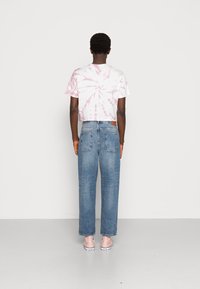 Rosa tie-dye t-shirt med kort ärm, lösa blå denimjeans, rosa slip-on-skor, stående mot en neutral bakgrund.