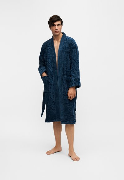 Homme debout pieds nus portant un peignoir texturé bleu foncé avec des poches et une ceinture sur un fond clair uni.