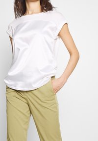 Mujer vistiendo una blusa blanca de manga corta con frente de satén y pantalones beige, de pie con una mano en el bolsillo frente a un fondo liso.