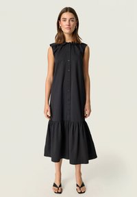 Vestido negro sin mangas con un frente abotonado, detalle de hombros con volantes y un dobladillo en capas. Confeccionado en una tela suave con una silueta fluida.