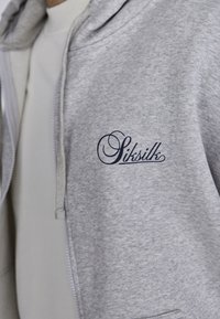 Sivá mikina na zips s jemnou textúrou, nastaviteľnou šnúrkou a tmavomodrým vyšitým logom "Siksilk" na ľavej hrudi.