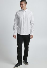 Chemise blanche à manches longues, fabriquée en tissu léger, associée à un pantalon noir et des baskets noires.