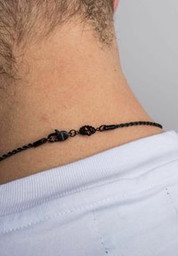 Musta punottu ketju, jossa on kynnen muotoinen kiinnitys ja lootuksen muotoinen yksityiskohta, joka on käytössä paljaalla iholla valkoisen paidan alla kaulalla.