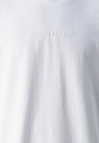 Tricou alb din bumbac, cu guler rotund și logo-ul embosat "GUESS JEANS" în centru. Textură moale cu un finisaj neted.