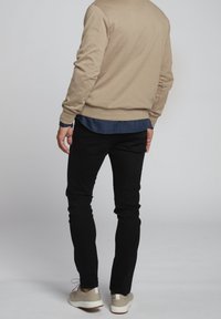Homme debout, dos tourné vers la caméra, portant un pull beige, une chemise bleue, un pantalon noir et des baskets beiges sur un fond gris clair uni.