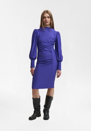 Purple fitted dress with puff sleeves and side ruching, made of a smooth fabric. Paired with black knee-high boots.