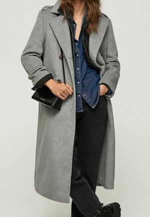 Kvinde iført en lang grå frakke over en denimskjorte og sorte bukser, holder en lille sort taske med den ene hånd i lommen.