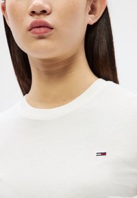 Tuvplāns personai ar taisniem tumšiem matiem, kas valkā baltu kreklu ar mazu sarkanu, baltu un zilu taisnstūra logotipu krūtīs.