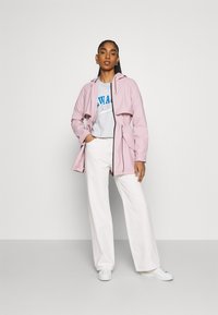 Casaco impermeável rosa claro com cintura ajustável, combinado com uma t-shirt gráfica cinza e calças largas brancas. A modelo usa ténis brancos.