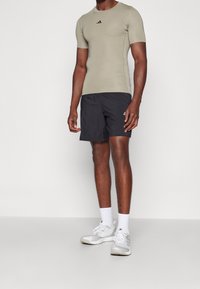 Strój sportowy składający się z dopasowanej, krótkorękawowej oliwkowej koszulki i czarnych szortów, zestawiony z białymi skarpetami i szarymi butami sportowymi.