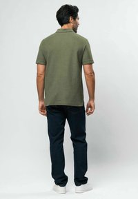 Hombre de pie de espaldas, vistiendo una camisa tipo polo de manga corta color verde oliva, jeans azul oscuro y zapatillas blancas sobre un fondo liso.