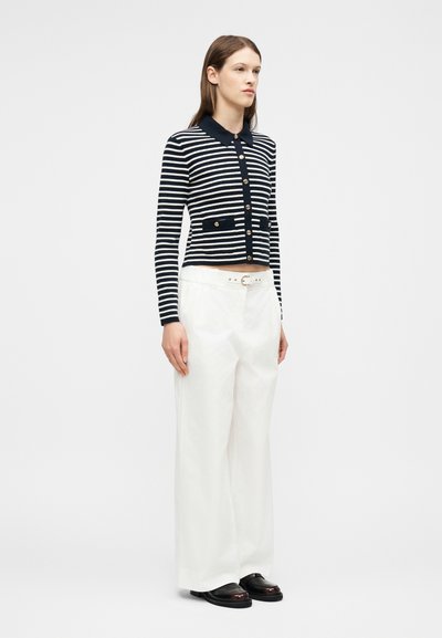 Femme debout portant un cardigan boutonné à rayures noires et blanches, un pantalon large blanc avec ceinture, et des chaussures noires, sur un fond uni.
