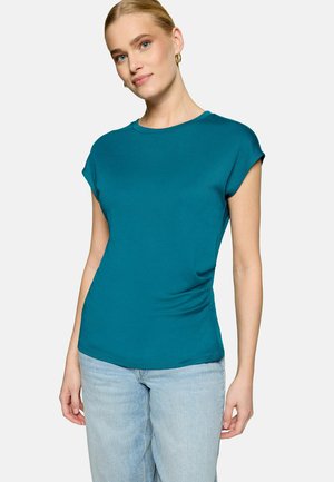 Femme portant un top sans manches couleur sarcelle et un jean bleu clair, debout devant un fond clair uni, regardant légèrement sur le côté.