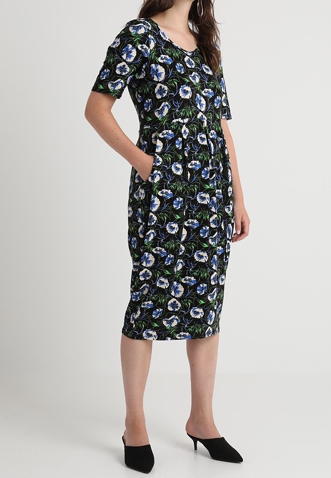 Robe noire à fleurs avec des manches courtes, présentant des motifs floraux bleus et blancs, une longueur jusqu'aux genoux et des poches latérales. Portée avec des chaussures noires.