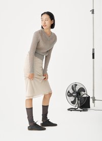 Γυναίκα με γκρι πουλόβερ και μπεζ φούστα σηκώνει το τελείωμα της φούστας κοντά σε έναν μαύρο ανεμιστήρα δαπέδου σε λευκό στούντιο.