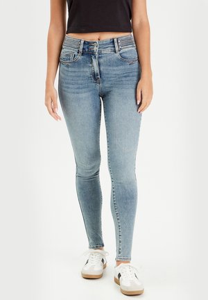 Kvinde iført lyseblå højtaljede skinny jeans med dobbelt knaplukning, kombineret med hvide sneakers og sort croppet top.
