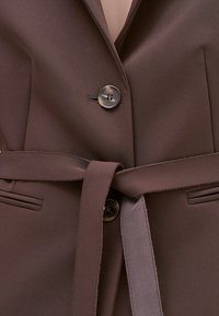 Brązowa wytworna marynarka z dopasowanym paskiem związanym w talii, z zapięciem na dwa guziki i rozciętymi kieszeniami po obu stronach.