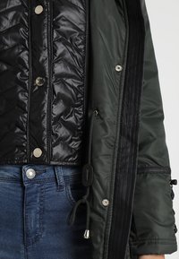 Veste matelassée noire avec boutons-pression argentés portée sous une veste vert foncé avec cordon de serrage, associée à un jean en denim bleu.