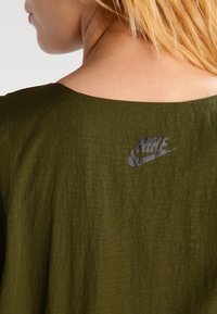 Top de tela texturizada en verde oliva con un escote amplio y un logo de Nike negro visible en la parte posterior. Diseño casual, material ligero.