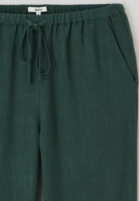 Pantalon en lin vert foncé avec une taille élastique, un cordon de serrage et des poches latérales. La texture est lisse et légère.