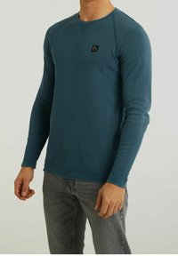 Man wearing teal textured long-sleeve shirt and gray jeans, standing against a plain light background.