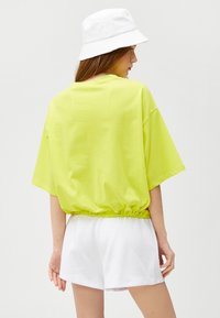 Neon grøn oversized t-shirt med elastik i kanten, sammen med hvide shorts og en hvid bøttehat. Glat bomulds materiale, afslappet pasform.