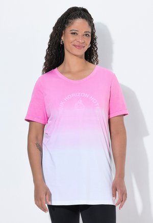 Kobieta z długimi kręconymi włosami, ubrana w różowo-biały gradientowy T-shirt z podwiniętymi rękawami i czarne spodnie, uśmiechająca się i stojąca przy białej ścianie.