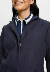 Giacca blu navy con texture, colletto e chiusura a un bottone. Sotto, una maglia blu navy con zip, dotata di una cerniera e rifiniture del colletto di colore crema.