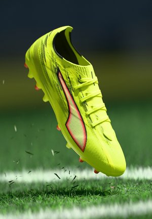 Lime green football boot with textured upper, three laces, red accents, and orange studs, suspended above grassy field with scattered debris.