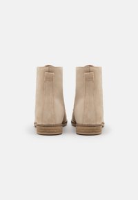 Bottines beige en daim à texture lisse, avec un bout arrondi et un talon empilé. Présentent un design minimaliste avec une languette de tirage à l'arrière.