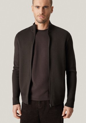 Mężczyzna ubrany w ciemnobrązowy kardigan na zamek, pod którym ma pasującą brązową koszulkę i sztruksowe spodnie, stoi na jasnym tle.