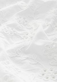 Tissu blanc ornée d'une broderie florale complexe avec une texture en relief, mettant en valeur des points surélevés et un tombé doux.