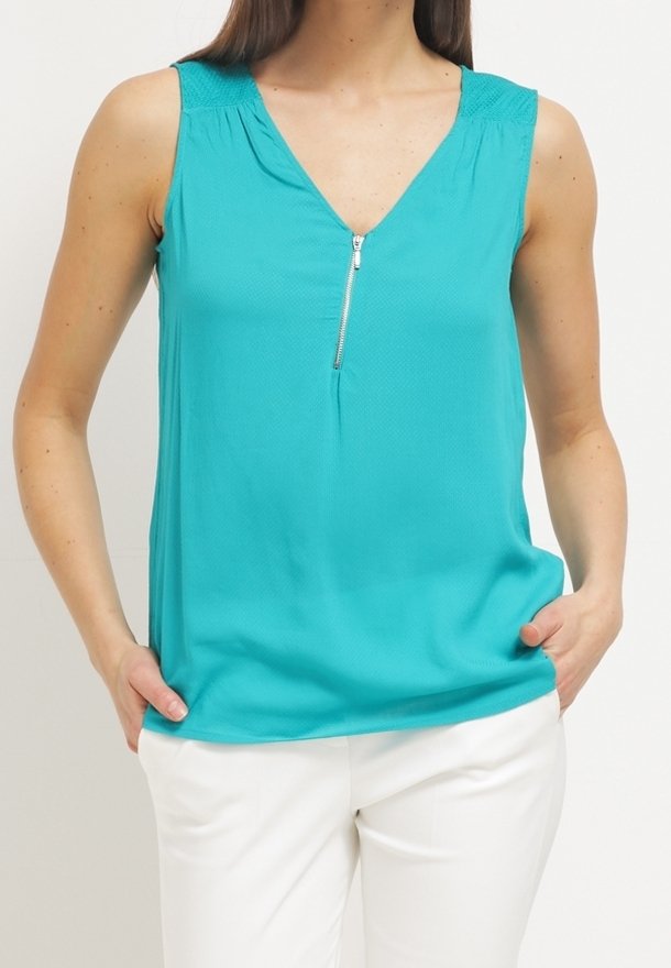 Haut turquoise sans manches avec col en V zippé et pantalon blanc, porté par une personne les mains dans les poches.