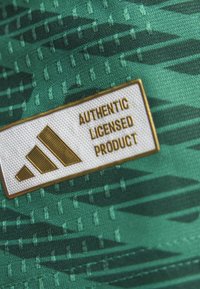 Zelená tkanina s texturovaným vzorem; obsahuje bílou nášivku s nápisem "AUTENTICKÝ LICENCOVANÝ PRODUKT" a zlatým logem Adidas.