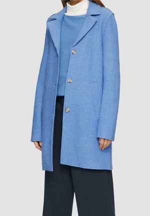 Femme portant un manteau bleu clair jusqu'aux genoux avec trois boutons, par-dessus un pull bleu et un col roulé blanc, associé à un pantalon noir à jambes larges.