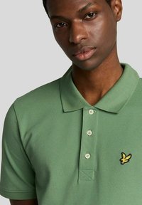 Polo verde realizzata in tessuto a trama, con colletto, tre bottoni e un logo ricamato giallo sul petto a sinistra.