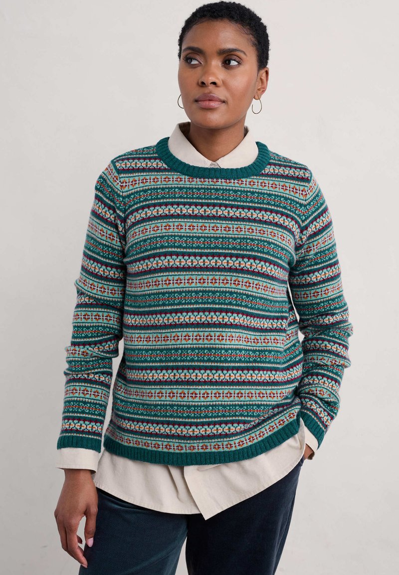 Gebreide trui in het groen met meerkleurige geometrische patronen, geribbelde hals en gelaagd met een witte blouse eronder.