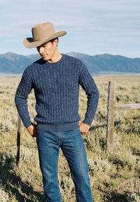 Homme en pull bleu, jean et chapeau de cowboy beige debout près d'une clôture en fil de fer dans un champ herbeux avec des montagnes en arrière-plan.