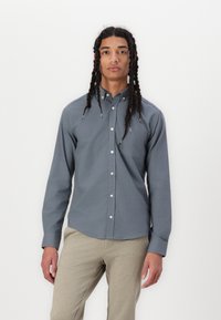 Camisa de botonadura en tela gris oscuro, de mangas largas, con un solo bolsillo en el pecho, dobladillo redondeado, combinada con pantalones beige claros.