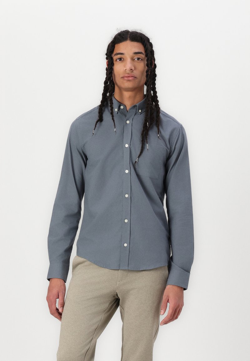 Camisa de botonadura en tela gris oscuro, de mangas largas, con un solo bolsillo en el pecho, dobladillo redondeado, combinada con pantalones beige claros.