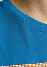 T-shirt turchese con collo rotondo testurizzato, caratterizzato da una striscia orizzontale a pois giallo-marrone nella parte inferiore.