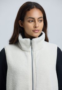 Witte fleece vest met hoge kraag, voorzien van een rits aan de voorkant en een gestructureerd oppervlak. Draagt over een zwart longsleeve shirt.
