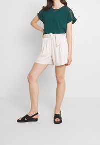 Chemise à manches courtes bleu sarcelle avec accents aux épaules, associée à un short beige taille haute avec ceinture, et des sandales noires à brides.