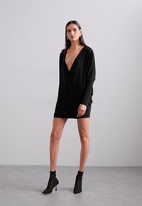 Zwarte wikkeljurk met een diepe V-hals, losse mouwen en een getailleerd silhouet. Gepaard met doorschijnende zwarte enkellaarsjes en minimalistische sieraden.