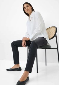 Witte overhemd met knoopdetails, gecombineerd met een donkere spijkerbroek en zwarte platte schoenen. Model zit op een zwarte metalen stoel met een geweven rugleuning.