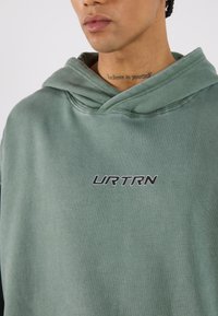 Felpa verde con cappuccio e logo frontale che recita "URTRN" in nero. Il tessuto è morbido, con un design rilassato e dettagli di cucitura visibili.